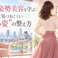 大阪城を望む明るく上品な部屋で、白いトップスとピンクのスカート姿の女性が鏡で後ろ姿を確認し、大阪の姿勢美容で背中や立ち姿を整える様子を示している。